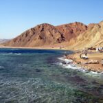 A praia do Blue Hole: os cafés beduínos ao pé das montanhas ocre, alguns carros e o mar agitado em primeiro plano