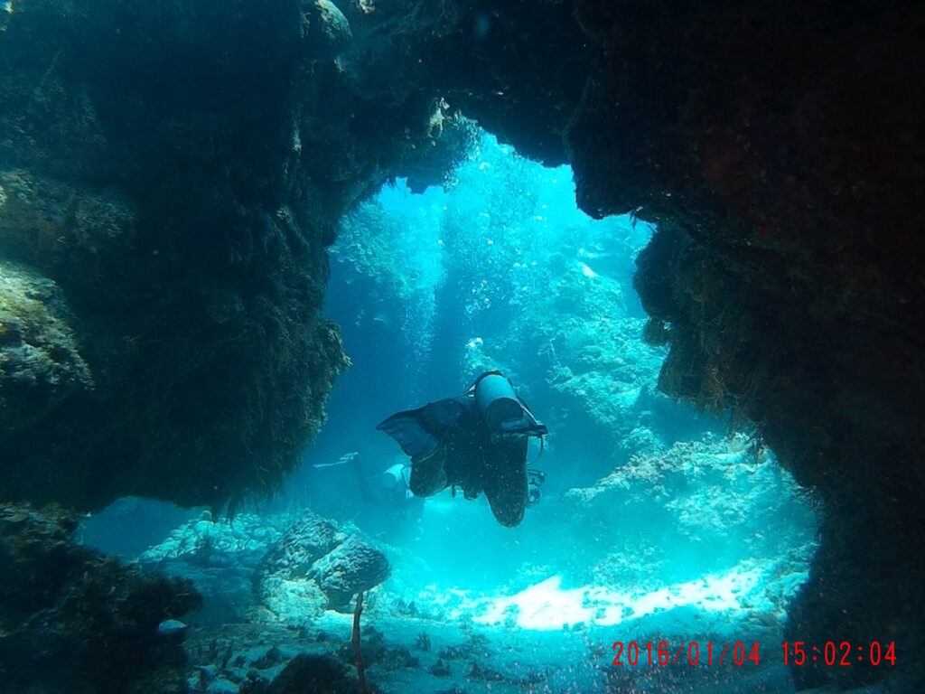 Um mergulhador atravessa um arco de coral escuro em Palancar, com a areia branca iluminada pelo sol atrás dele