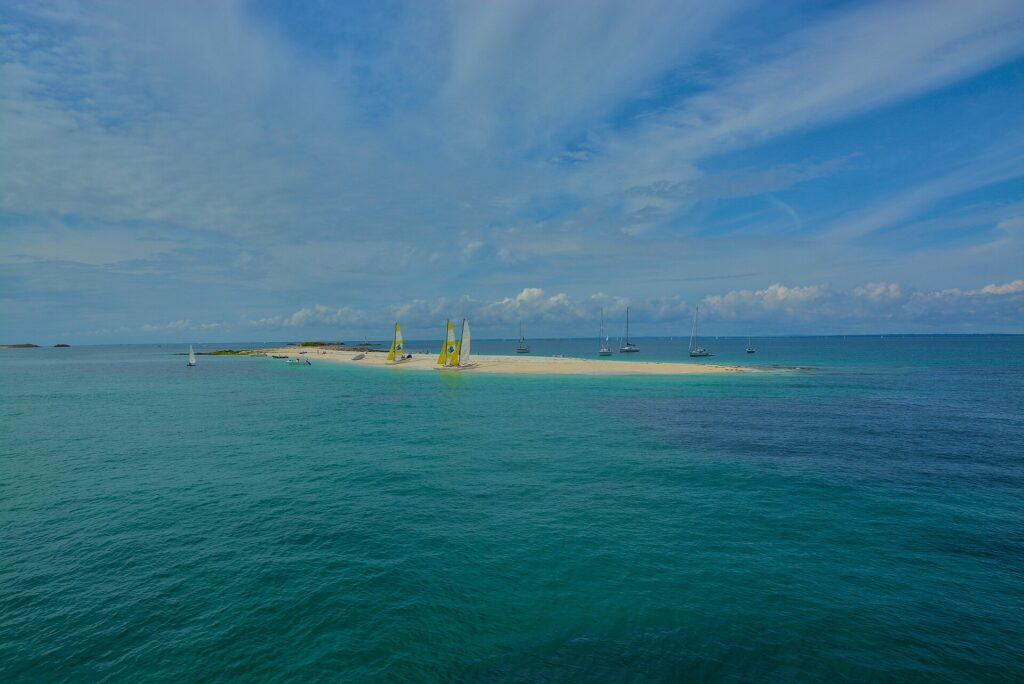 Uma longa língua de areia branca onde estão encalhados catamarãs de velas amarelas e brancas, cercada de veleiros ancorados numa água turquesa, sob um céu azul riscado de nuvens.