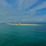 Uma longa língua de areia branca onde estão encalhados catamarãs de velas amarelas e brancas, cercada de veleiros ancorados numa água turquesa, sob um céu azul riscado de nuvens.