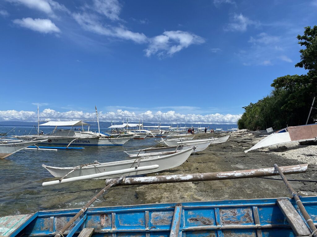 Uma fila de bangkas brancas amarradas ao longo de uma costa rochosa, pequenas canoas com flutuadores puxadas sobre a rocha e um barco azul descascado em primeiro plano, sob um céu azul com nuvens brancas.