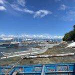 Uma fila de bangkas brancas amarradas ao longo de uma costa rochosa, pequenas canoas com flutuadores puxadas sobre a rocha e um barco azul descascado em primeiro plano, sob um céu azul com nuvens brancas.