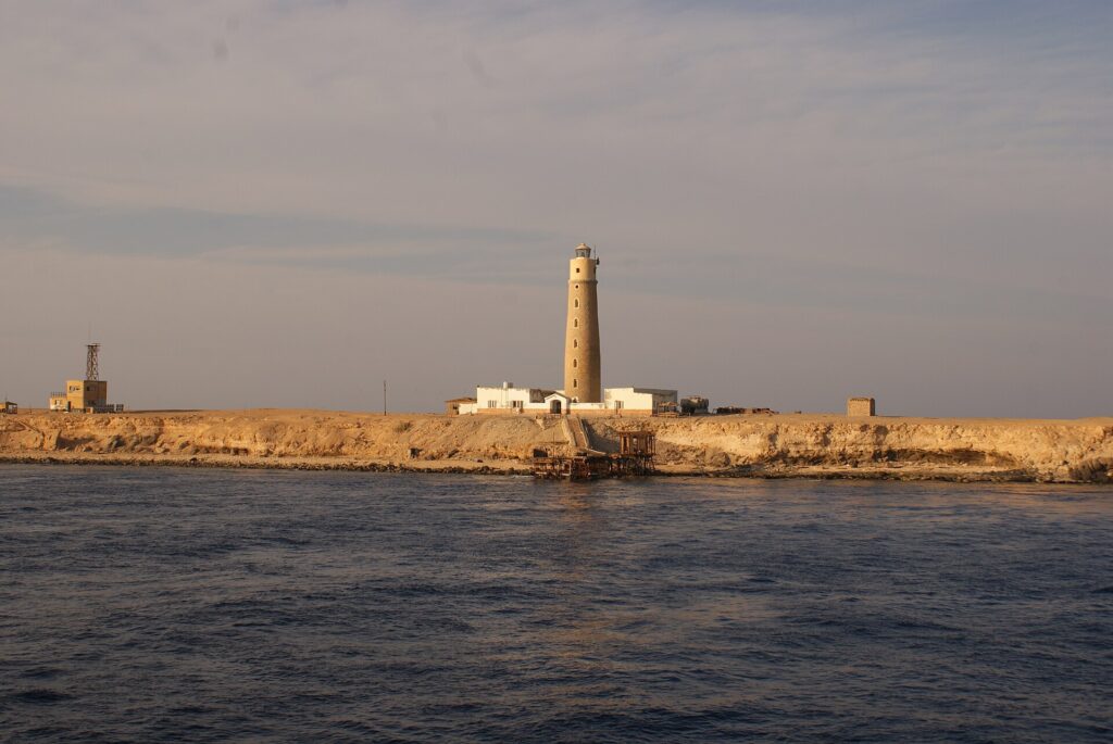 O farol de Big Brother e seus prédios brancos na ilhota desértica, com um píer enferrujado em primeiro plano, na luz dourada da tarde