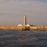 O farol de Big Brother e seus prédios brancos na ilhota desértica, com um píer enferrujado em primeiro plano, na luz dourada da tarde