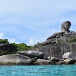 O enorme bloco de granito equilibrado da ilha 8 coroa um amontoado de rochas cinza e lisas à beira de uma água turquesa, sob um céu azul enevoado, com uma linha de boias em primeiro plano