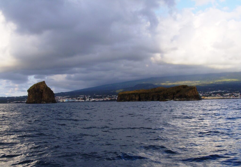 Vistas de um barco num mar escuro e agitado, uma ilhota rochosa erguida à esquerda e uma ilhota longa e plana à direita, diante da cidade de Madalena e das encostas do Pico sob um céu carregado