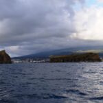 Vistas de um barco num mar escuro e agitado, uma ilhota rochosa erguida à esquerda e uma ilhota longa e plana à direita, diante da cidade de Madalena e das encostas do Pico sob um céu carregado
