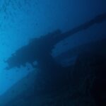 O canhão de popa do Thistlegorm em silhueta contra o azul, sob um cardume de peixinhos