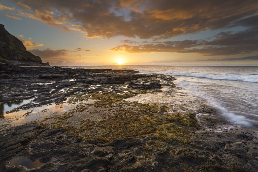 Pôr do sol sobre o oceano visto de um platô de basalto preto coberto de algas e poças, com as ondas lambendo à direita e um penhasco à esquerda