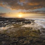 Pôr do sol sobre o oceano visto de um platô de basalto preto coberto de algas e poças, com as ondas lambendo à direita e um penhasco à esquerda