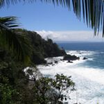 Vista do alto emoldurada por folhas de palmeira: um cabo coberto de floresta tropical desce até o mar, as ondas se desfazem em espuma branca sobre rochas negras e uma costa distante se desenha no horizonte sob um céu azul.