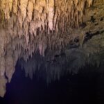Um teto de estalactites bege iluminado por lanterna, refletido numa superfície de água negra, no cenote The Pit
