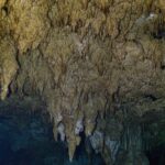 Uma floresta de estalactites bege que desce em direção à água escura, com uma coluna no centro e um brilho verde à direita