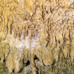 Close de formações amarelo-claras e estalactites finas, com o reflexo ondulado da água embaixo