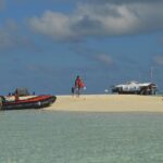 Um banco de areia branca cercado de água clara verde-pálida, onde um bote inflável preto e laranja acaba de atracar, com pessoas de colete salva-vidas na areia e a estação dos guardas sobre palafitas à direita.