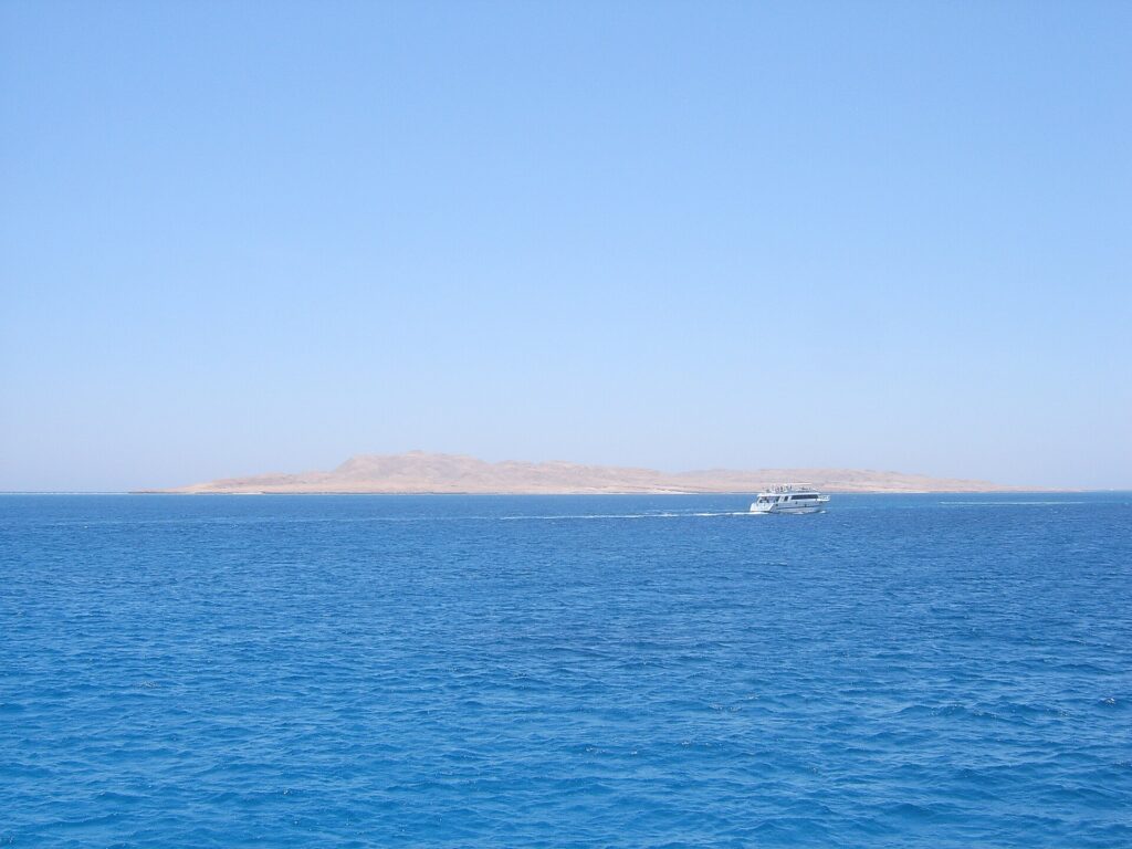 A ilha plana e desértica de Giftun Soraya vista do mar, com um barco de mergulho branco passando na frente
