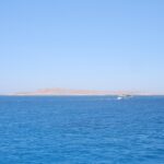 A ilha plana e desértica de Giftun Soraya vista do mar, com um barco de mergulho branco passando na frente