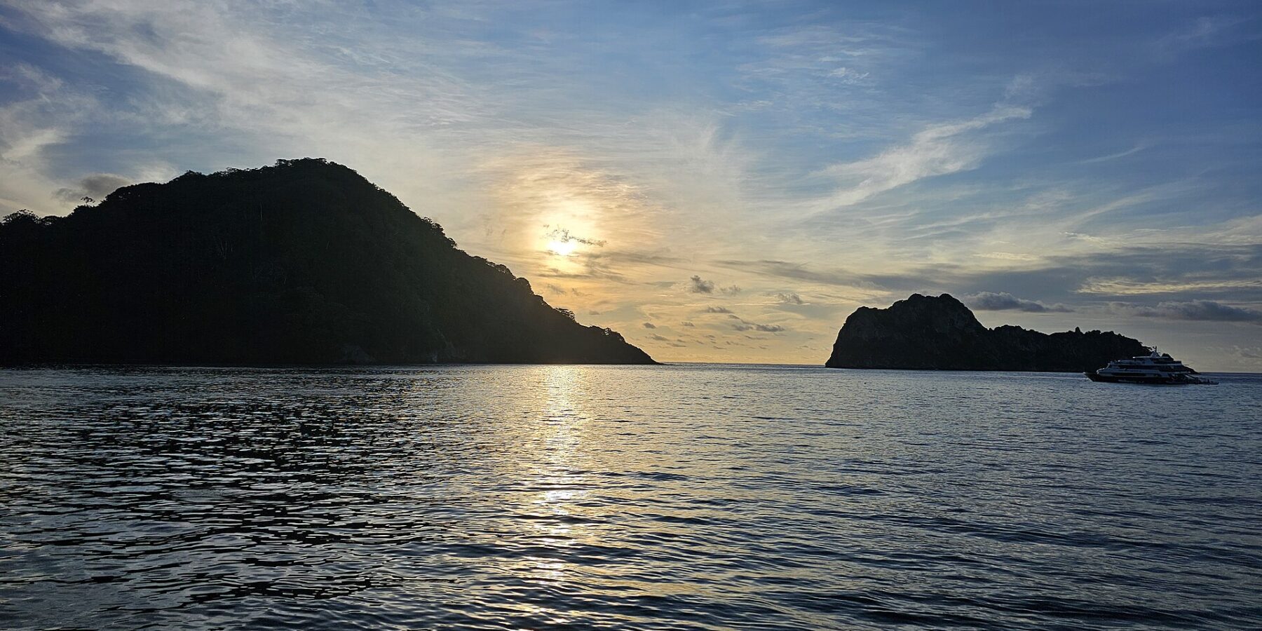A Ilha do Coco contra a luz ao pôr do sol, com um barco de cruzeiro ancorado à direita e o mar calmo