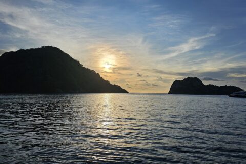 A Ilha do Coco contra a luz ao pôr do sol, com um barco de cruzeiro ancorado à direita e o mar calmo