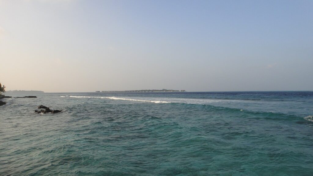 De uma margem rochosa, um canal turquesa agitado onde uma linha de ondas quebra sobre o recife e, no horizonte, uma ilha baixa margeada de bangalôs sobre palafitas, sob um céu pálido.