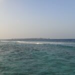 De uma margem rochosa, um canal turquesa agitado onde uma linha de ondas quebra sobre o recife e, no horizonte, uma ilha baixa margeada de bangalôs sobre palafitas, sob um céu pálido.