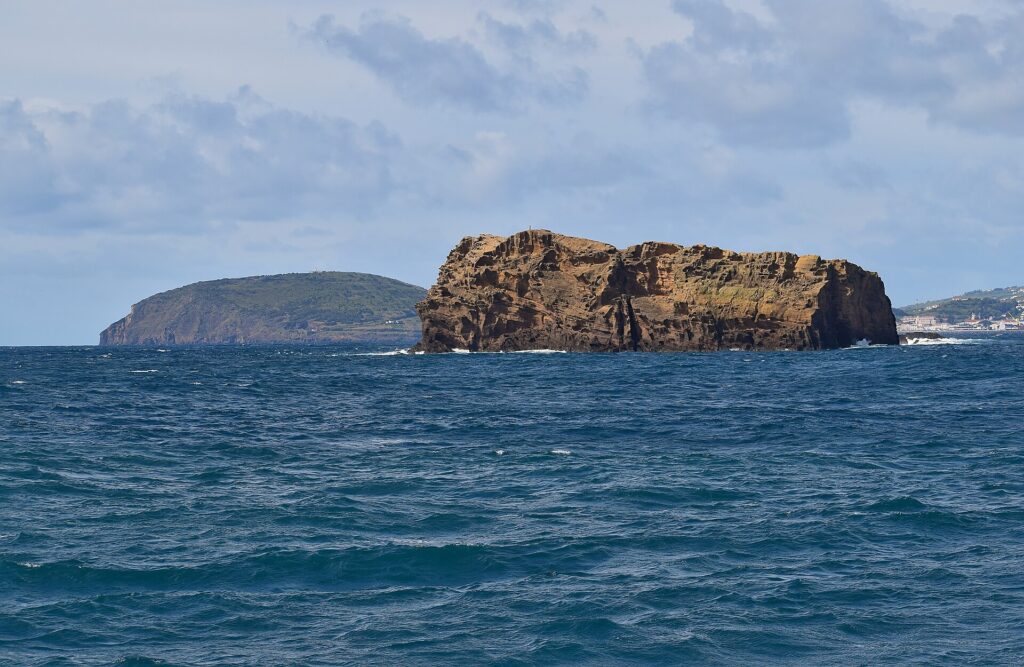 O Ilhéu Deitado, longo bloco de tufo ocre com falésias cheias de alvéolos, num mar azul-escuro agitado, com a colina verde do Monte da Guia atrás e casas do Faial ao longe, à direita