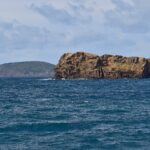 O Ilhéu Deitado, longo bloco de tufo ocre com falésias cheias de alvéolos, num mar azul-escuro agitado, com a colina verde do Monte da Guia atrás e casas do Faial ao longe, à direita