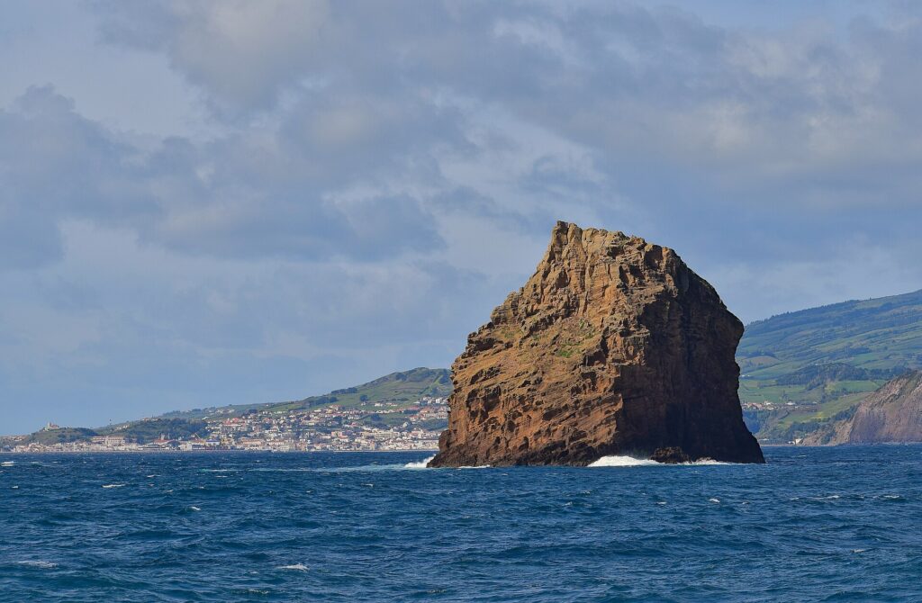 O Ilhéu em Pé, alto rochedo de tufo ocre com camadas escavadas, se ergue num mar azul-escuro onde quebram algumas ondas, diante da cidade branca da Horta e das colinas verdes do Faial sob um céu nublado