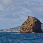 O Ilhéu em Pé, alto rochedo de tufo ocre com camadas escavadas, se ergue num mar azul-escuro onde quebram algumas ondas, diante da cidade branca da Horta e das colinas verdes do Faial sob um céu nublado