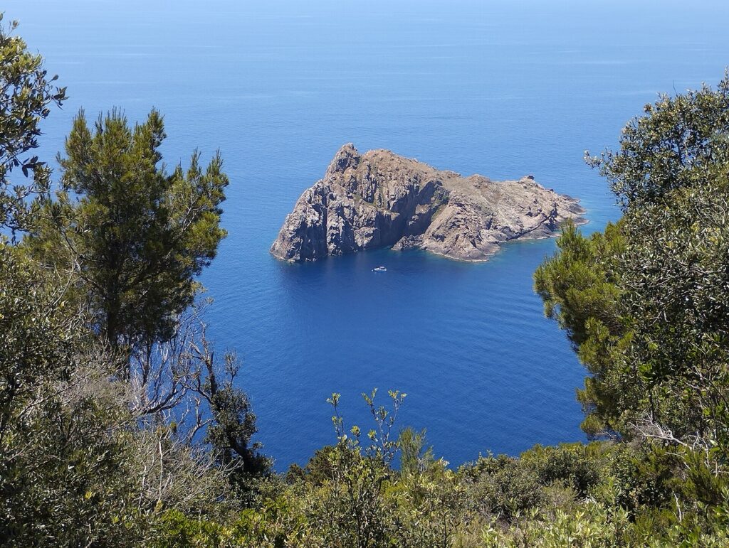 Vista do alto de Port-Cros, entre pinheiros e arbustos, sobre a ilhota rochosa e escarpada de La Gabinière num mar azul profundo, com um pequeno barco branco ancorado ao pé dela.