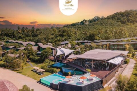 Vista aérea do Kalima Resort Khao Lak ao pôr do sol, com piscinas e telhados curvos ao pé de uma colina arborizada