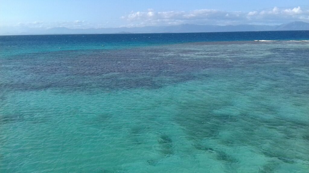 A lagoa vista do alto, água turquesa salpicada de blocos de coral escuros, a barreira onde as ondas quebram à direita e as montanhas da Grande Terre no horizonte