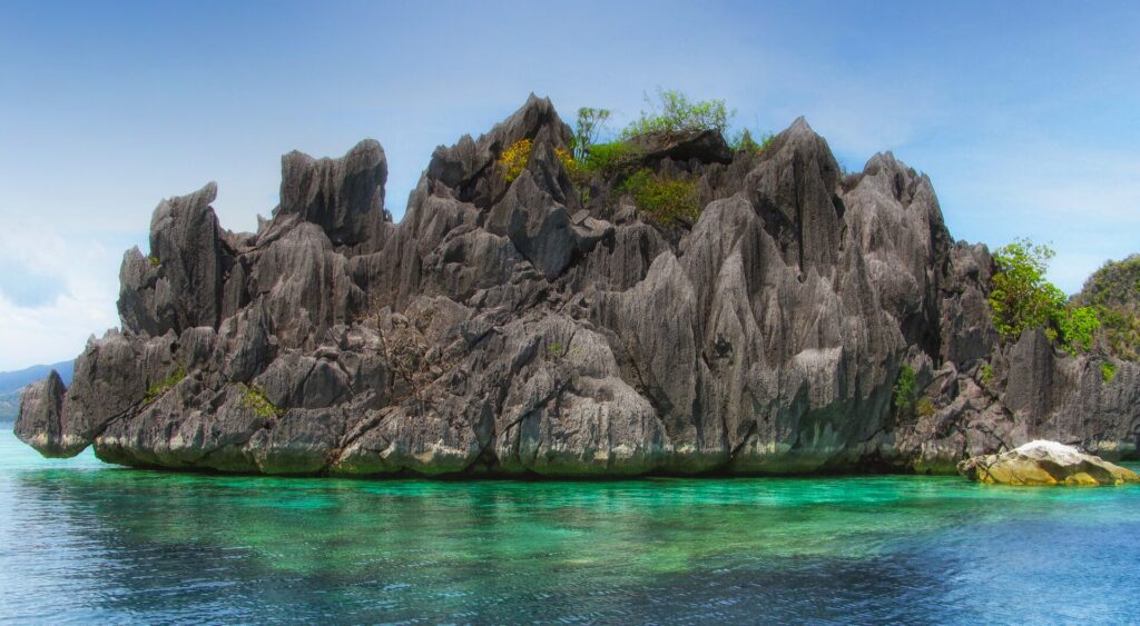 Uma ilhota de calcário cinza cheia de pontas, coberta por um pouco de vegetação, se ergue acima de uma água turquesa transparente onde se adivinha o coral, sob um céu azul.