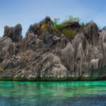 Uma ilhota de calcário cinza cheia de pontas, coberta por um pouco de vegetação, se ergue acima de uma água turquesa transparente onde se adivinha o coral, sob um céu azul.