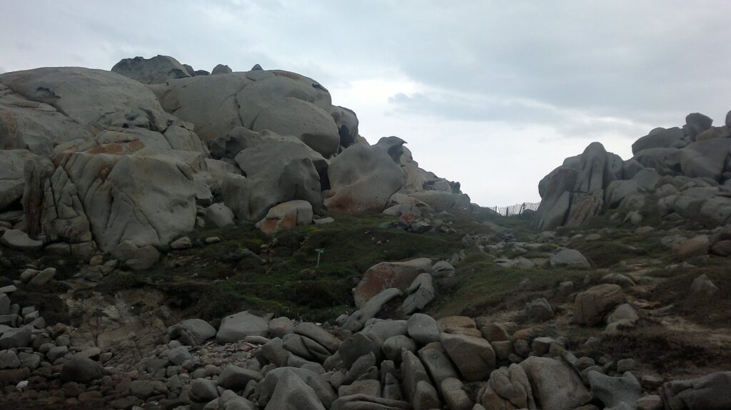 Amontoado de grandes blocos de granito cinza arredondados e escavados pela erosão, manchados de líquens laranja, ao redor de um gramado rasteiro onde uma trilha margeada por uma cerca de madeira segue para o mar, sob um céu nublado.