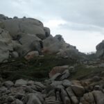 Amontoado de grandes blocos de granito cinza arredondados e escavados pela erosão, manchados de líquens laranja, ao redor de um gramado rasteiro onde uma trilha margeada por uma cerca de madeira segue para o mar, sob um céu nublado.