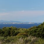 Vista do mato da costa sobre uma longa ilha baixa feita de blocos de granito claro, num mar azul-escuro, com um veleiro ao pé dela e montanhas azuladas no horizonte.