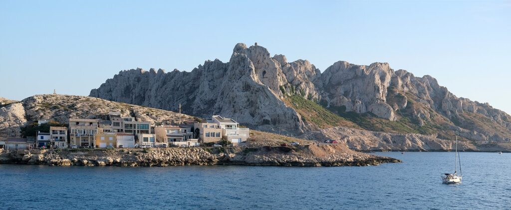 As casas da vila de Les Goudes alinhadas rente a uma costa de rochas, diante de uma alta crista de calcário branco com picos recortados, sob um céu azul, com um veleiro ancorado num mar calmo.