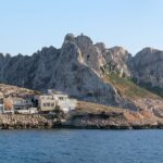 As casas da vila de Les Goudes alinhadas rente a uma costa de rochas, diante de uma alta crista de calcário branco com picos recortados, sob um céu azul, com um veleiro ancorado num mar calmo.