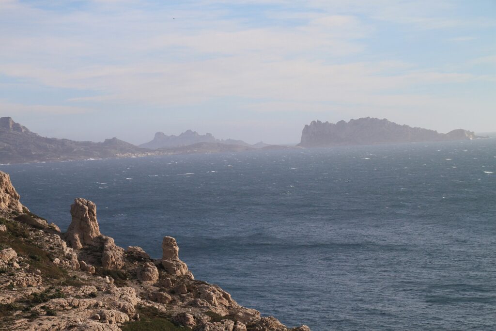 Das rochas calcárias do Frioul, em primeiro plano, vista de um mar agitado e cheio de espuma até a ilha Maïre e a costa de Les Goudes, recortadas na bruma.