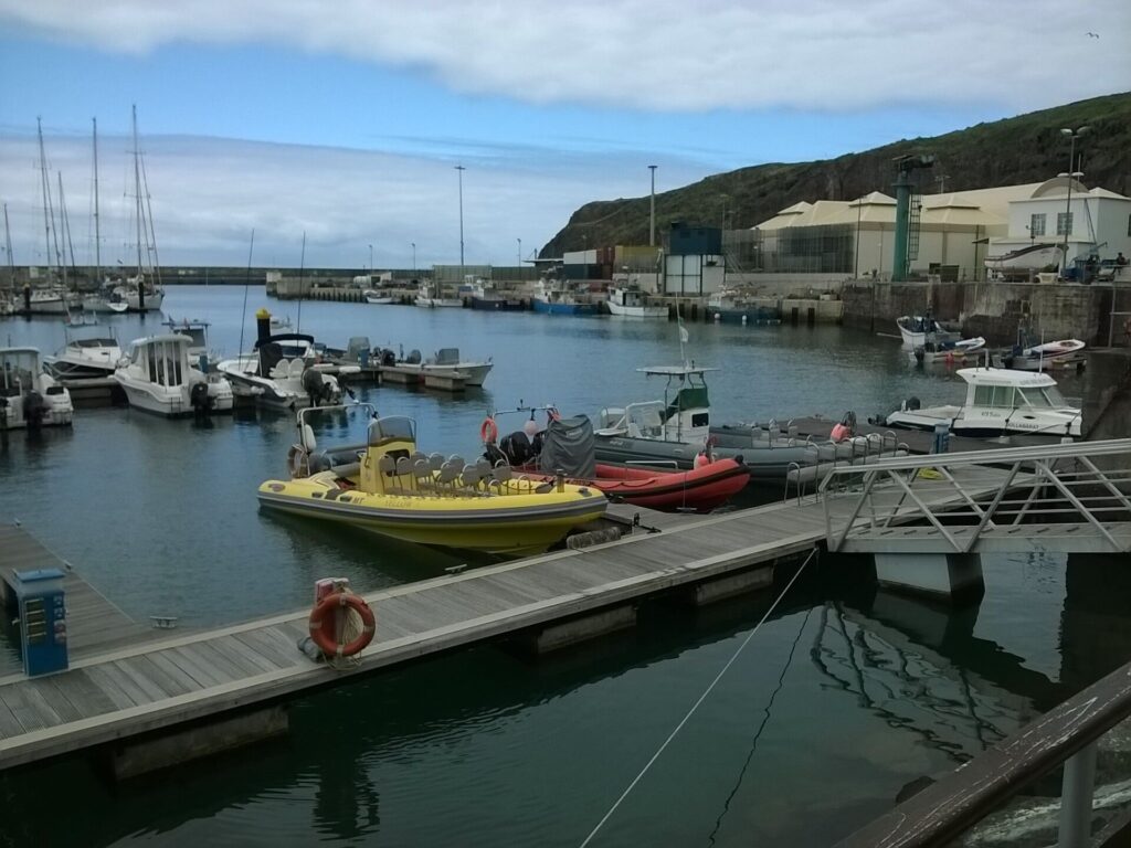 Botes semirrígidos, um amarelo com fileiras de assentos e um vermelho, atracados no píer da marina de Vila do Porto, com veleiros e barcos de pesca ao fundo sob um penhasco verde e um céu nublado