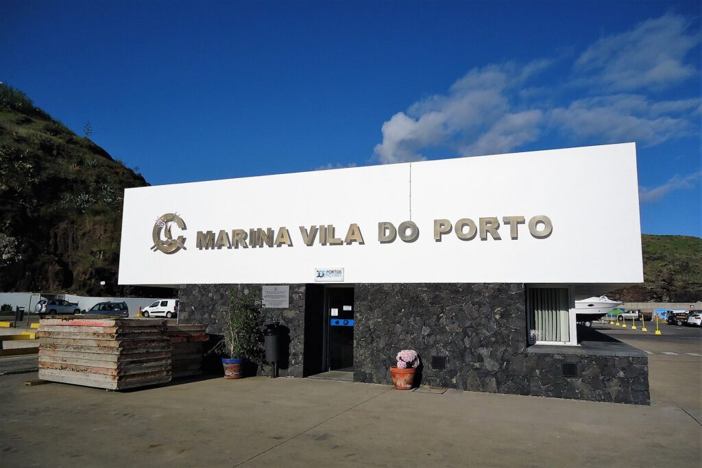 O prédio de recepção da marina de Vila do Porto, grande letreiro branco com letras metálicas sobre um muro de basalto preto, sob um céu azul, com um penhasco verde à esquerda