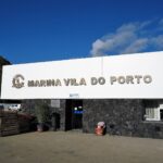 O prédio de recepção da marina de Vila do Porto, grande letreiro branco com letras metálicas sobre um muro de basalto preto, sob um céu azul, com um penhasco verde à esquerda