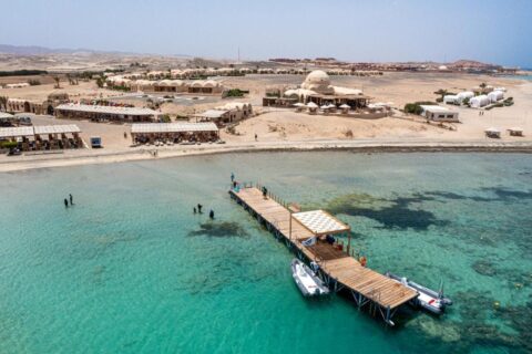Vista aérea do Marsa Shagra Village, com o píer, dois botes infláveis e a vila à beira da água turquesa