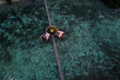 Vista aérea noturna de um longo píer de madeira que sai da praia, com duas cabanas sobre palafitas no meio do caminho