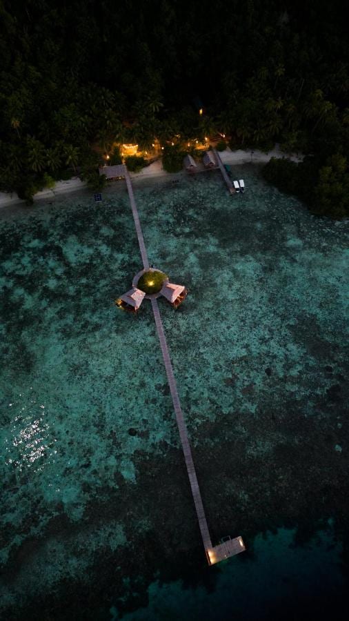 Vista aérea noturna de um longo píer de madeira que sai da praia, com duas cabanas sobre palafitas no meio do caminho