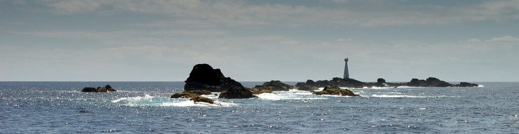 Panorama da cadeia de escolhos pretos das Formigas rente à água, orlados de espuma branca num mar azul cintilante, com o farol branco de Formigão à direita sob um céu velado