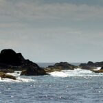 Panorama da cadeia de escolhos pretos das Formigas rente à água, orlados de espuma branca num mar azul cintilante, com o farol branco de Formigão à direita sob um céu velado