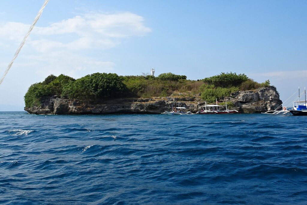 A ilha de Pescador vista do mar, coberta de arbustos verdes, com o farol branco no alto e três bancas de mergulho amarradas ao pé da falésia