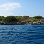 A ilha de Pescador vista do mar, coberta de arbustos verdes, com o farol branco no alto e três bancas de mergulho amarradas ao pé da falésia